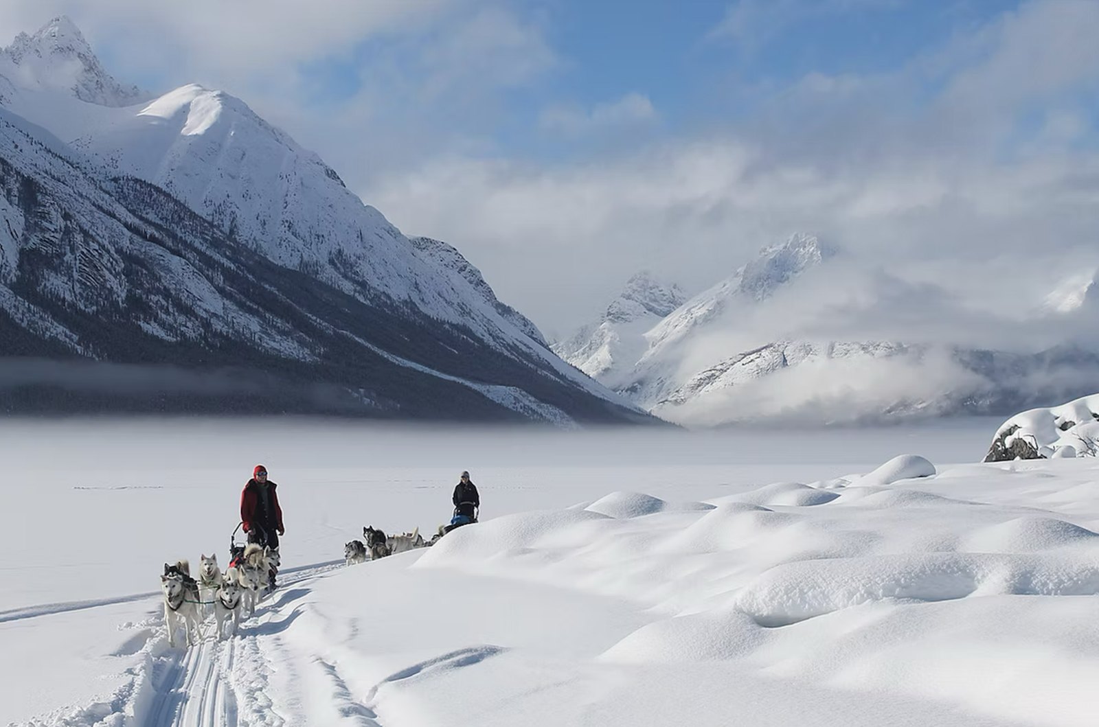 Two dog teams on a frozen lake below snowy mountains
